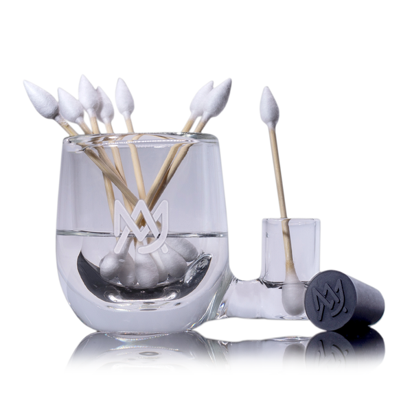 Glass holder displaying white cotton swabs with wooden sticks standing upright, featuring a logo with interlocking "M" and "J" on a smooth surface, surrounded by a reflective white background.