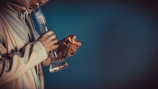 Person lighting a glass bong for smoking, wearing a hooded jacket in a dimly lit setting